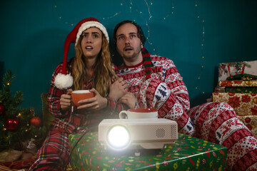 Young couple getting scared while they watch horror movie on the projector with Christmas pijama.
