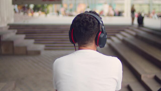 A young handsome man in a white T-shirt and headphones sits on the steps in the city and listens to music on the phone or watches a video