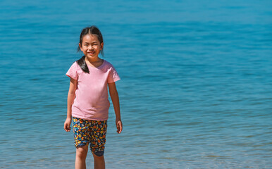 Fototapeta na wymiar Portrait Asian child girl standing on beach under sunny sky, background blurred sea. Asian child with smiling face, looking at camera, wearing casual, black long hair. Space for copy and design.