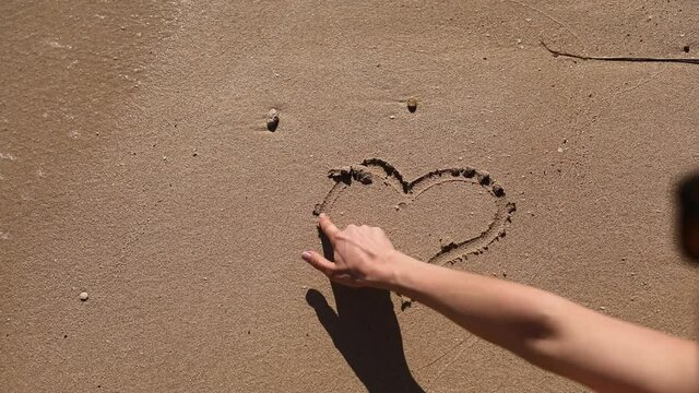 Woman draws heart on a wet sand. Female's hands writing the word sea on sand. Sea water washes away the drawing on a sandy beach