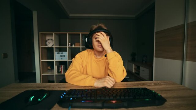 An emotional gamer in a headset plays a video game on a computer at home and loses with a sad face and removes the headphones from his head. The guy lost in an online computer game.