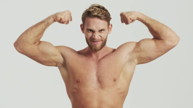 A strong half-naked blonde man is showing his muscles standing isolated over a grey background in the studio