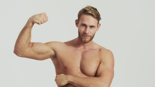 A smiling half-naked blonde man is showing his biceps muscles standing isolated over a grey background in the studio
