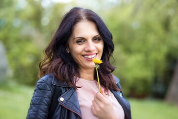 Middle-age woman in black leather jacket smiling, smelling yellow flower.