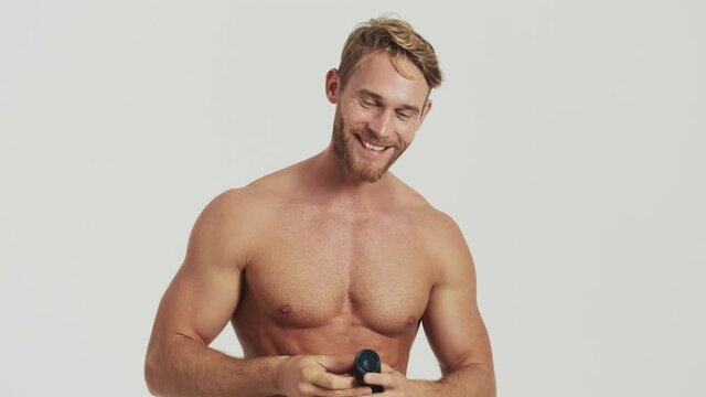 A happy half-naked blonde man is using deodorant spay standing isolated over a grey background in the studio