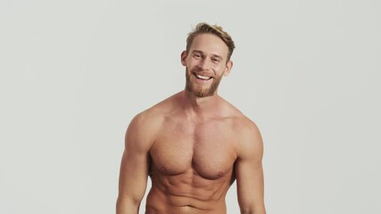 A smiling half-naked blonde man is showing his pectoral muscles posing to the camera standing isolated over a grey background in the studio