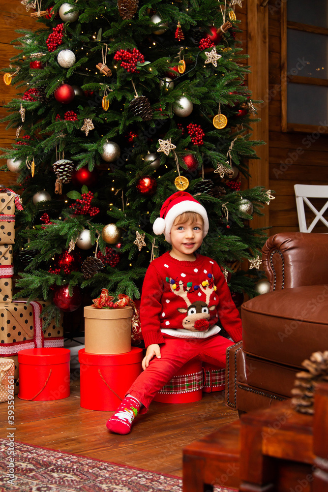 Poster a boy in a New Year's sweater and a santa claus hat under a big tree looks at gifts