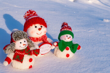 Three toy snowmen in hats and scarves in the rays of light on the snow smile and wish merry Christmas