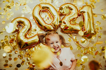 Cheerful child girl with numbers 2021 rejoices in golden confetti flying from above, top view. Merry Christmas and New Year celebration concept. Holidays at home