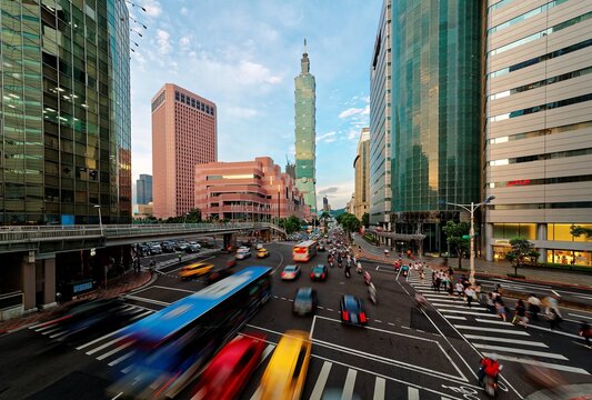 View of a busy street corner in Downtown Taipei City at rush hour with cars & buses dashing by, Taipei 101 Tower & World Trade Center Building in Xinyi Financial District & people passing on crosswalk