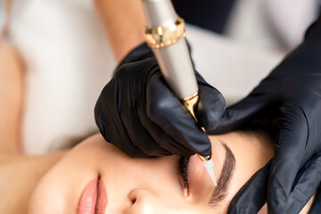 Beautician applying permanent makeup on eyebrows of young woman by special tattoo machine tool