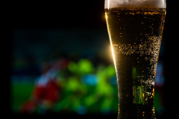 Close-up of fresh foaming light beer in a glass and football on TV on background. Sports bar concept