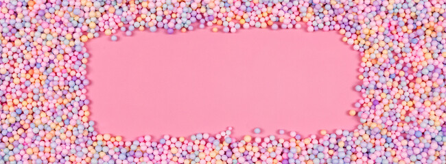 Frame made of pastel color balls on pink. Styrofoam or Polystyrene foam background. Mix of colorful sugar balls for sweets decoration. Rainbow colored sugar chips, dragee spins, confectionery.