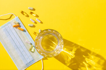 Protective face masks, medicinal pills and a glass of water on a yellow background with sunlight shadow.