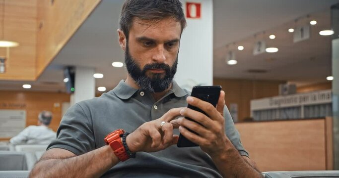 Handsome bearded man texting message on mobile phone while sitting at hospital lobby waiting doctor visit. Young man using virtual assistant app to sets reminder