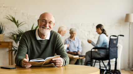 Senior man in eyeglasses smiling happily at camera while reading book sitting at table in assisted...