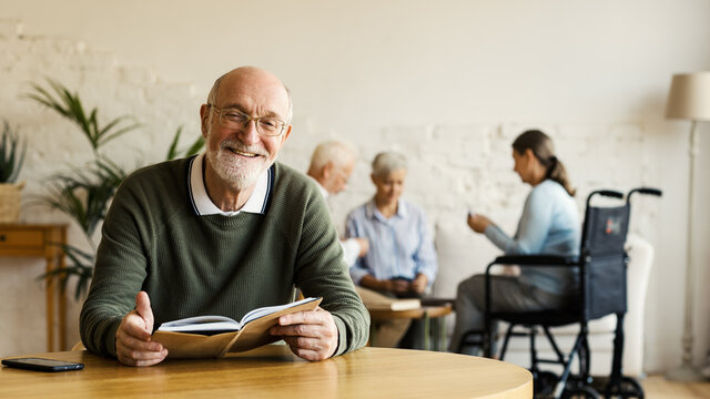 Senior man in eyeglasses smiling happily at camera while reading book sitting at table in assisted living home. Three elderly people including disabled woman playing cards in background