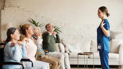 Side view of young nurse and group of senior patients including disabled ones doing arm rotation exercise together in nursing home