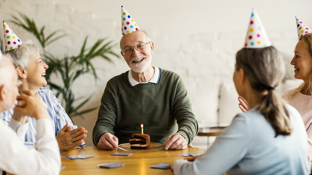 Elderly birthday man smiling happily after receving birthday cake with burning candle while playing cards with aged friends in party hats in nursing home