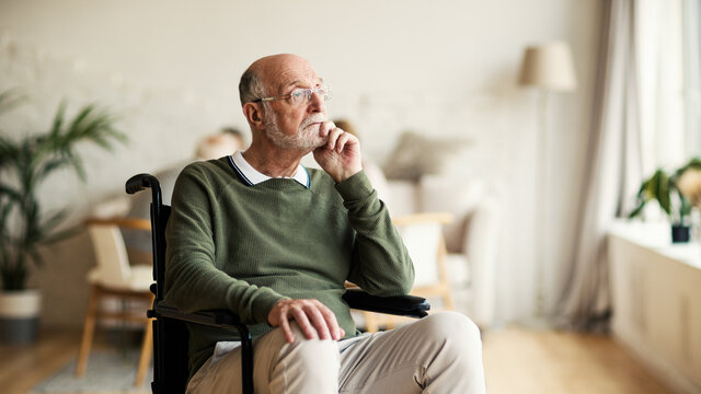 Disabled senior man in eyeglasses sitting in wheelchair looking away and thinking with his hand on chin in nursing home