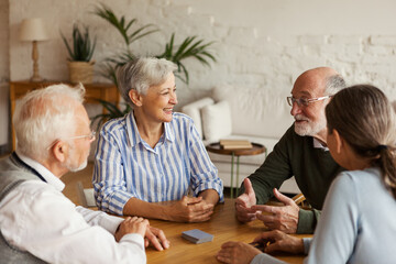 Group of four cheerful senior friends, two men and two women, sitting at table and enjoying talk after playing cards in assisted living home - obrazy, fototapety, plakaty