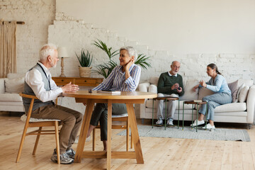 Senior man and woman sitting at table and enjoying talk, another aged couple interacting in background sitting on sofa in common room of nursing home - Powered by Adobe
