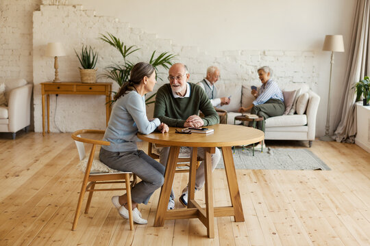 Senior man and woman sitting at table and talking, another aged couple playing cards in background sitting on sofa in common room of assisted living home