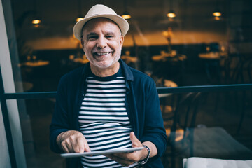 Portrait of happy Caucaisan blogger on retirement holding digital tablet in hands and smiling at camera during rest daytime in city, cheerful male senior in straw hat using touch pad on leisure