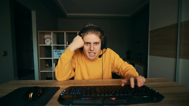 Dissatisfied gamer in a headset plays video games at home on the computer, looks at the camera and gets angry. An upset gamer makes a streaming game.