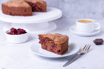 Chocolate cherry pie on a white plate. In the background is a pie, a cup of coffee, cherries and pieces of chocolate.