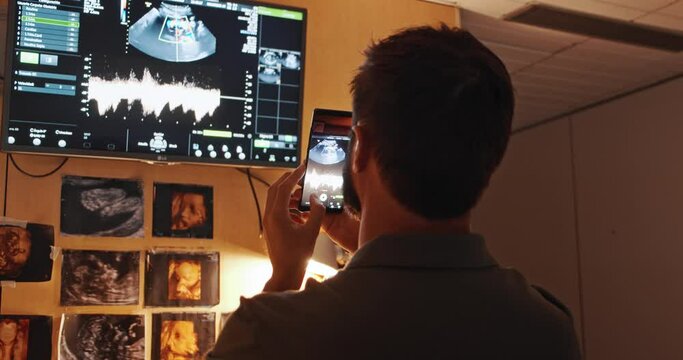 Young man taking mobile photo and video of ultrasound his future child in sonography cabinet. Carried ultrasound of a pregnant woman in the 6th month of the baby