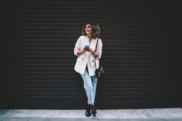 Funny female with mobile phone in hands rejoicing near mock up wall with copy space area for text advertising, cheerful hipster blogger with modern cellphone feeling excited at publicity area