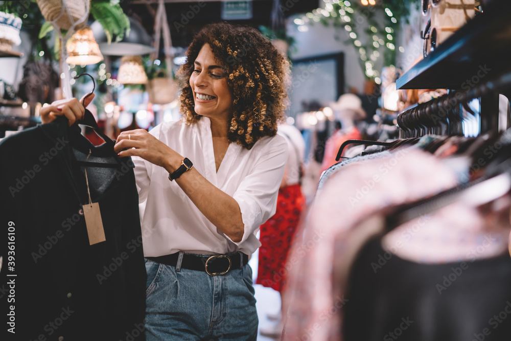 Wall mural Cheerful female standing near hangers with brand clothes and laughing during Black Friday shopping with discounts, happy hipster girl enjoying pastime for update her wardrobe spending day in boutique