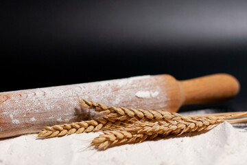 A kitchen rolling pin, spikelets of wheat, and a stack of white flour lie on a dark surface.