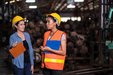 Engineer woman wearing helmet standing with friend at the warehouse where keep machine parts and holding clipboard. Used vehicle parts for recycling in the scrap yard garage. Reuse and recycle concept