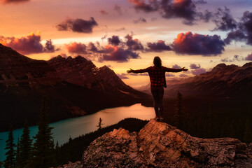 Adventurous girl with open arms standing on the edge of a cliff overlooking the beautiful Canadian Rockies and Peyto Lake. Dramatic, vibrant summer sunset. Banff National Park, Alberta, Canada.