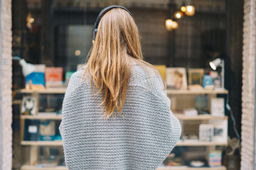 Young blonde woman on her back with headphones and pullover looking at a bookstore window.