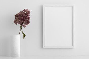 Empty white frame on a white wall. Dry decorative branch in a vase on a shelf or table. White color. Clean, modern, minimal frame, the layout of the poster. Internal interior.