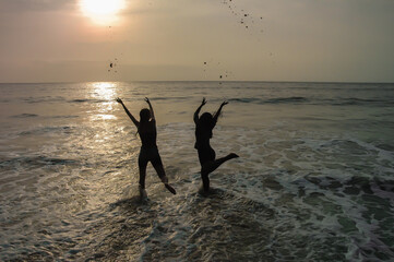 Silhouette of two happy women on african beach throwing sand in the air at sunset.