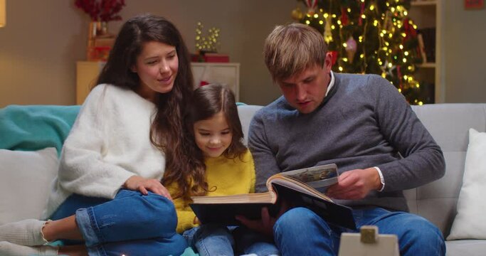Portrait of joyful Caucasian litle kid girl sitting at cozy decorated room with mom and dad in good mood reading book together on Christmas Eve. Happy holidays. New Year celebration concept