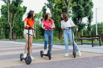 Fototapeta premium E-scooter, electric scooter, Ecological urban city transport, alternative energy, alternative lifestyle. Three Young girl friends on vacation having fun driving electric scooter through city park