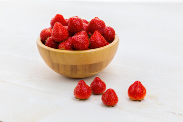 Full wooden bowl of fresh strawberries on white table. Red raw berries rich in vitamin inside wooden dish with few others on side. Sweet healthy fruit on desk.