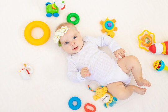 small baby girl 6 months old lying on her back on a white bed at home among toys, top view