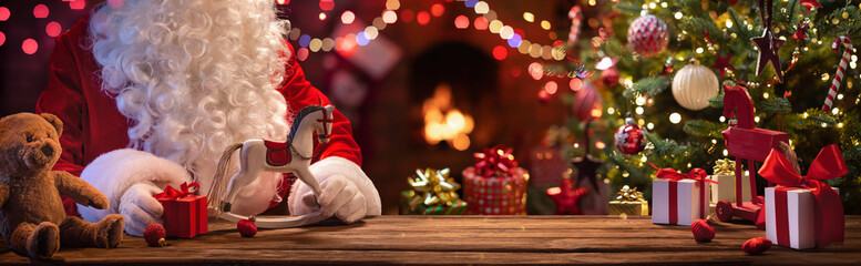 Santa Claus Sits At A Table With Gifts And Toys Near The Fireplace And Christmas Tree - obrazy, fototapety, plakaty