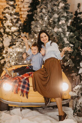 Mother and son sit on car hood and have a fun against background of Christmas tree.