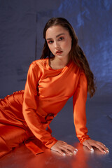 Studio portrait. Elegant woman sitting and posing in orange silk dress against blue metaallic background. High fashion female model with make-up and wet hairstyle