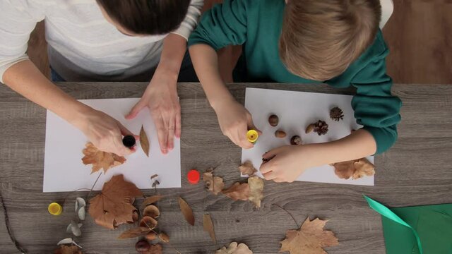 family, creativity and craft concept - mother and little son with glue sticks and paper making pictures of dry autumn leaves, pine cones and chestnuts at home