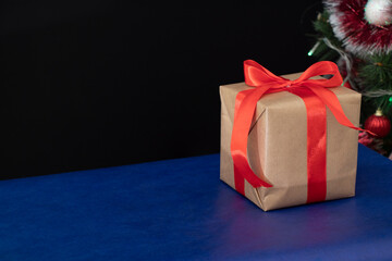 A gift wrapped in craft paper with a red wide ribbon stands on a blue table near the Christmas tree. Holidays.