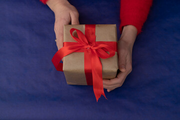 Gift wrapped in craft paper with a wide red ribbon in hands on a blue background. Holidays
