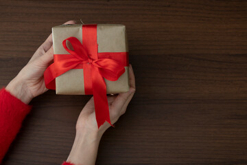 Female hands in a red sweater hold a gift wrapped in craft paper with a wide red ribbon on a wooden background.